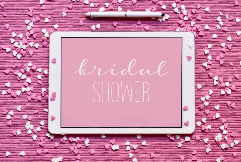 high-angle shot of a tablet with the text bridal shower written in white in it against a pink background, placed on a pink surface sprinkled with white and pink heart-shaped confetti sprinkles