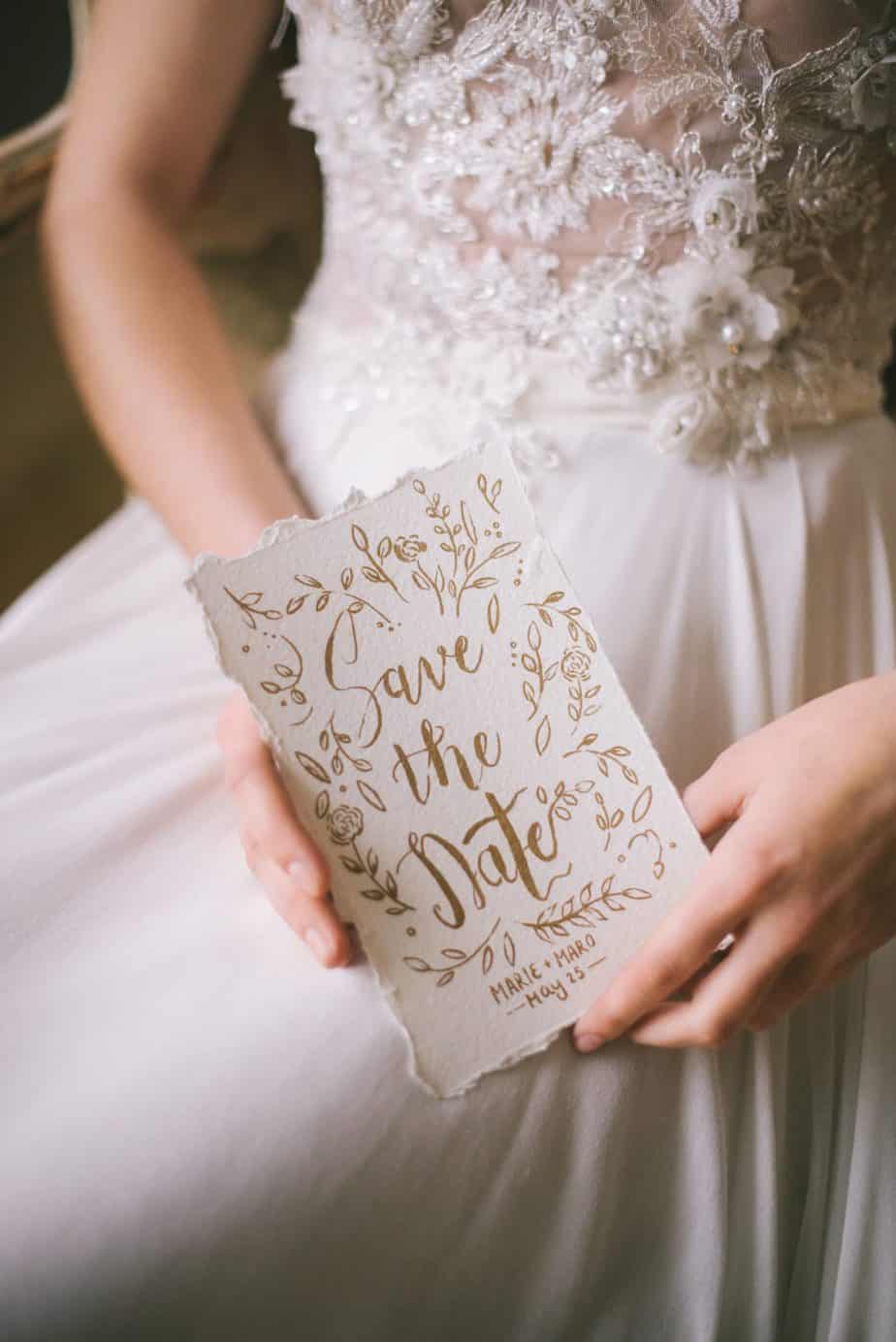 bride in white gown holding white and gold save the date for wedding