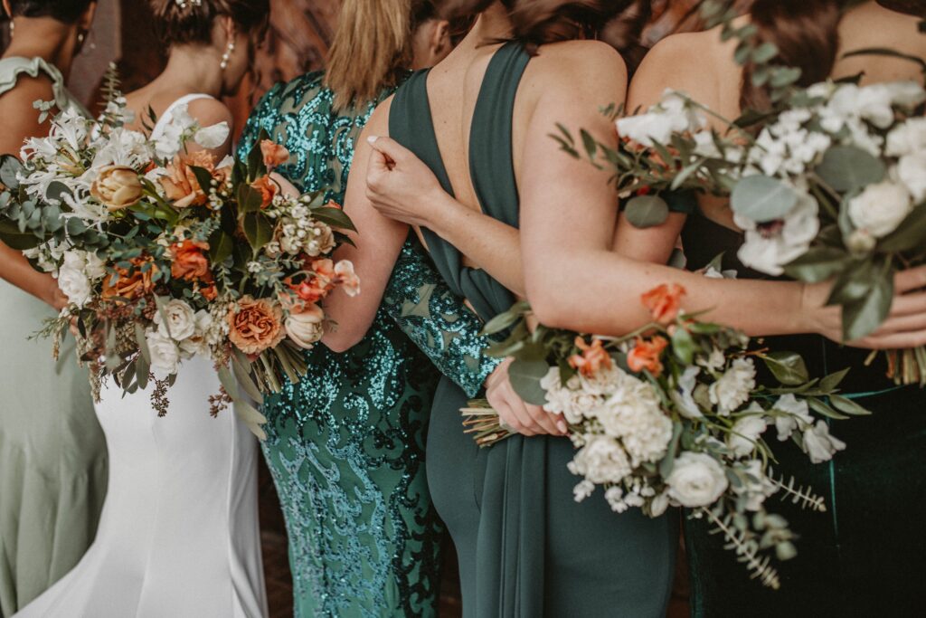 Photograph of 4 bridesmaids in mismatched emerald green dresses stand arm in arm with the bride to be with their backs facing the camera.